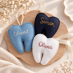 May include: Three plush tooth-shaped ornaments with hanging loops. One is light blue with the name Edward embroidered in white, one is navy blue with the name Simon in yellow, and one is white with the name Olivia embroidered in dark red.