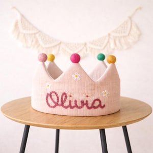 May include: A light pink fabric crown with colorful pom-poms and embroidered details. The crown has the name "Olivia" embroidered in a cursive font. Small white flower details are also embroidered on the crown. The crown is sitting on a wooden table.