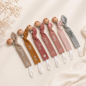 Puede incluir: Una colección de sujetachupetes para bebés en varios colores, incluyendo marrón, mostaza y rosa. Cada clip presenta un clip de madera y una correa de tela con un nombre bordado en blanco. Algunas correas tienen diseños bordados.