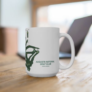 以下が含まれることがあります： 緑色のゴルフコースのアウトラインと「オーガスタ・ナショナル・ゴルフクラブ、オーガスタ、ジョージア州」というテキストが書かれた白いセラミックマグカップです。