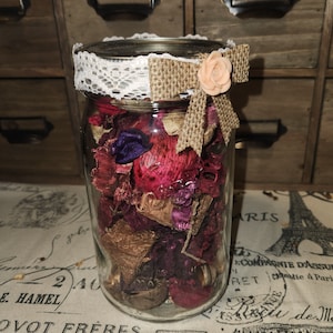 May include: A glass jar filled with dried flower petals in shades of pink, purple, and brown. The jar has a white lace and burlap bow with a pink flower accent.