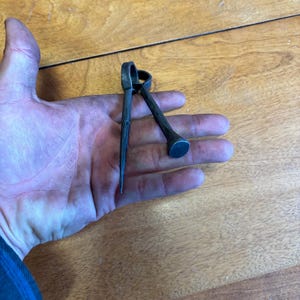 Puede incluir: Una herramienta de metal oscuro con un extremo puntiagudo y un extremo redondeado, sostenida en una mano. La herramienta tiene un anillo en la parte superior y el extremo puntiagudo es afilado. El extremo redondeado es plano. La herramienta parece hecha a mano.