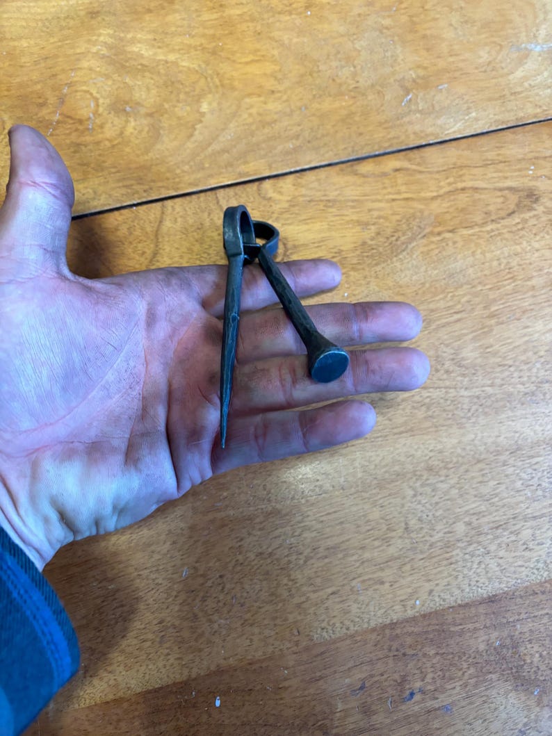 Puede incluir: Una herramienta de metal oscuro con un extremo puntiagudo y un extremo redondeado, sostenida en una mano. La herramienta tiene un anillo en la parte superior y el extremo puntiagudo es afilado. El extremo redondeado es plano. La herramienta parece hecha a mano.