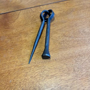 May include: Hand-forged metal tool with a heart-shaped handle. The tool has two arms, one pointed and the other with a flat, rounded head. The metal has a dark, textured finish. The tool is likely used for shaping or manipulating metal.
