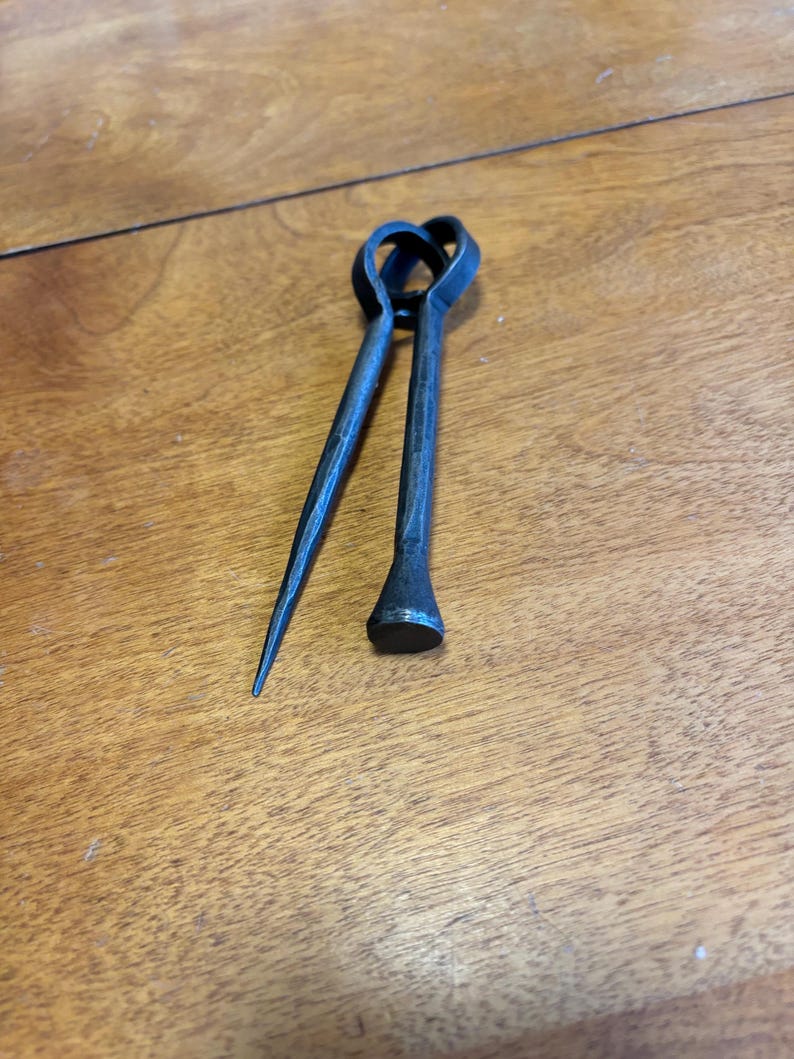 Puede incluir: Herramienta de metal forjada a mano con un mango en forma de coraz&oacute;n. La herramienta tiene dos brazos, uno puntiagudo y el otro con una cabeza plana y redondeada. El metal tiene un acabado oscuro y texturizado. La herramienta probablemente se utiliza para dar forma o manipular metal.