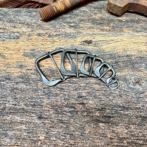 May include: A set of five graduated dark metal belt buckles arranged in a row, each featuring a rectangular frame and a central prong. The buckles are crafted from a dark, matte-finished metal and vary in size from largest to smallest.