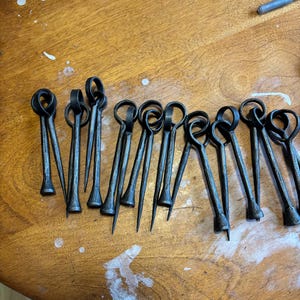 Puede incluir: Un conjunto de brochetas met&aacute;licas forjadas a mano, de color gris oscuro, con un lazo en la parte superior y una punta. Est&aacute;n dispuestas sobre una superficie de madera. El dise&ntilde;o sugiere su uso para asar o cocinar.