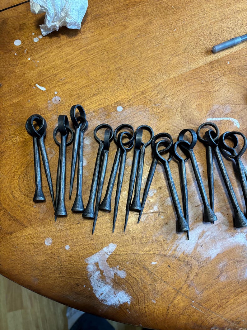 Puede incluir: Un conjunto de brochetas met&aacute;licas forjadas a mano, de color gris oscuro, con un lazo en la parte superior y una punta. Est&aacute;n dispuestas sobre una superficie de madera. El dise&ntilde;o sugiere su uso para asar o cocinar.