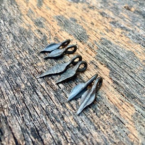 Puede incluir: Tres pares de pequeños dijes de metal forjados a mano, cada uno con forma de hoja puntiaguda o punta de lanza con un lazo circular en la parte superior. Los dijes tienen un acabado gris acero oscuro y texturizado, dispuestos en línea diagonal.