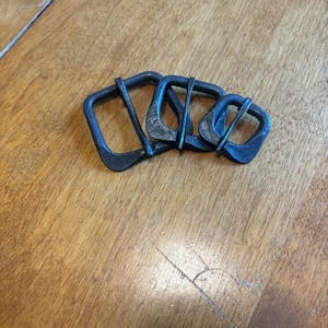 May include: Four dark metal buckles with a rectangular shape and a central bar. The buckles are arranged in a group on a wooden surface. The metal has a slightly textured appearance.