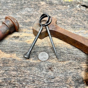May include: A hand-forged dark metal tool featuring two interlocking circular loops at the top, with one end tapering to a sharp point and the other end flared, positioned next to a United States quarter coin for scale on a weathered wood surface.