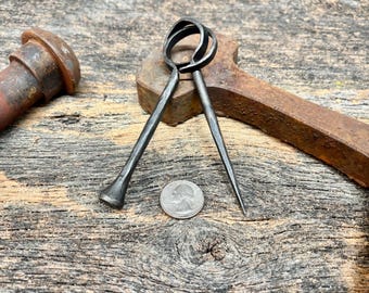 May include: A hand-forged dark metal tool featuring two interlocking circular loops at the top, with one end tapering to a sharp point and the other end flared, positioned next to a United States quarter coin for scale on a weathered wood surface.