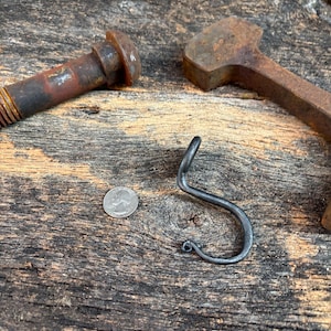 May include: A hand-forged dark gray metal S-hook with a curled end, positioned next to a silver-colored US quarter coin for scale. The hook features a smooth, curved design with a distinct loop at the bottom, showcasing a raw, industrial finish.