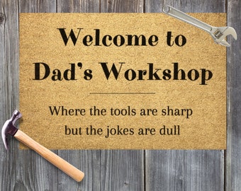 May include: A brown doormat with the text "Welcome to Dad's Workshop" and the phrase "Where the tools are sharp but the jokes are dull".