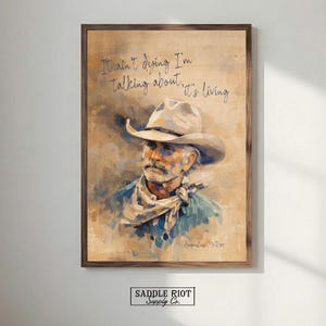 May include: Framed artwork featuring a portrait of a man in a cowboy hat and scarf. The artwork has a vintage aesthetic with the text "It ain't dying I'm talking about, it's living" in script. The artwork is in a wooden frame.