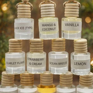 May include: A collection of small, clear glass bottles with wooden lids and hanging cords. Each bottle has a label with a different scent name, including "Black Ice," "Mango & Coconut," and "Vanilla." The bottles are arranged in a pyramid shape.