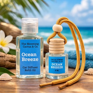 May include: Two clear glass car air freshener bottles with blue labels that read "Ocean Breeze." One bottle is a refill oil, the other is 8 ml. A yellow cord with a wooden bead and wooden discs is attached to the smaller bottle. The background is a beach scene.
