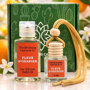 May include: Two clear glass bottles with red labels that read "FLEUR D'ORANGER" and "The Blooming Cactus & Co." are displayed. One is a car diffuser refill oil, and the other is a 10 mL bottle. A yellow rope and wooden diffuser are also visible, along with a green box.