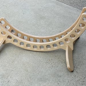 May include: A wooden stand with a curved shape and a series of holes. The stand is made of light wood and has a natural finish.