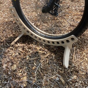 May include: A wooden bike stand with a cutout design to hold a bicycle wheel. The stand is made of light brown wood and has a natural finish. The stand is designed to hold a bicycle wheel with a disc brake.