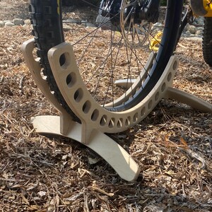 May include: A wooden bike stand with a curved design and multiple holes to hold a bicycle wheel. The stand is made of light brown wood and is resting on a bed of brown mulch.