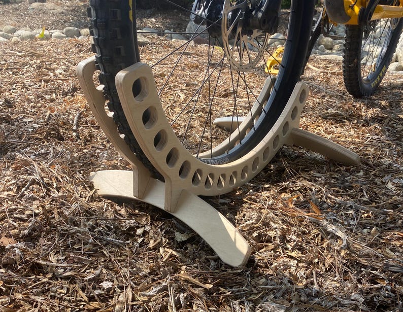 May include: A wooden bike stand with a curved design and multiple holes to hold a bicycle wheel. The stand is made of light brown wood and is resting on a bed of brown mulch.