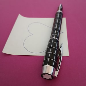 Könnte beinhalten: Ein schwarz-silberner Stift mit einem Gittermuster und einem silbernen Clip. Der Stift liegt auf einem Papier mit einem gezeichneten Herzen. Der Hintergrund ist magenta.
