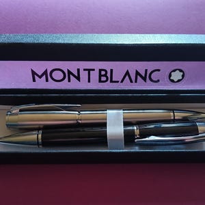 Peut inclure: Une boîte noire contenant un stylo argenté et un stylo noir. La boîte porte l'inscription "MONTBLANC".