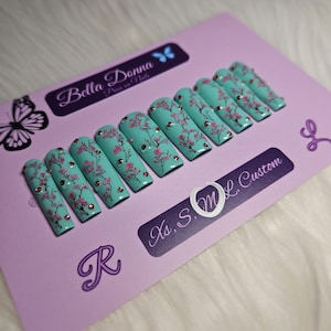 May include: A set of press-on nails in a light teal color, adorned with pink cherry blossom designs and small, sparkling embellishments. The nails are displayed on a lavender card with the text "Bella Donna Press on Nails" and "Xs, S, M, L Custom".