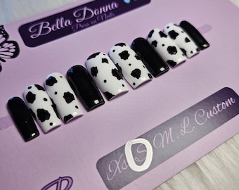 May include: A set of press-on nails in a cow print design, alternating black and white with black spots. The nails are displayed on a lavender surface with the text "Bella Donna Press on Nails" and "Xs O M. L. Custom".