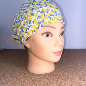 Puede incluir: Un gorro quirúrgico blanco con un estampado de limones amarillos y hojas azules. El gorro está atado en la parte posterior y está sobre una cabeza de maniquí. El texto "Modèle Citrons" es visible.