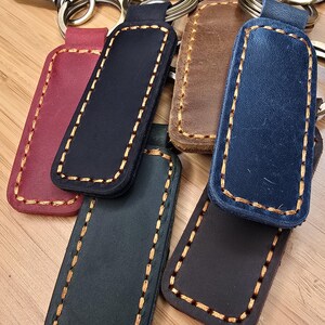 May include: A collection of rectangular leather keychains in various colors including burgundy, black, brown, and blue. Each keychain features thick, contrasting tan stitching along the edges and is attached to a circular metal split ring.