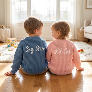 Könnte beinhalten: Ein blauer, langärmeliger Strickpullover mit weißem Sticktext Big Bro und ein rosafarbener, langärmeliger Strickpullover mit weißem Sticktext Lil Sis, beide mit gerippter Struktur und Rundhalsausschnitt, von hinten betrachtet.