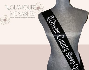 May include: Black satin sash with white text that reads "2023 Greene County Sheep Queen".