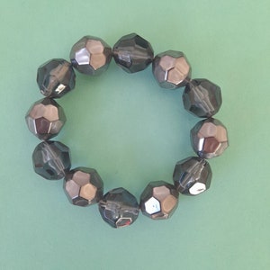 May include: A bracelet made of dark gray faceted beads. The beads are arranged in a circle and are connected by a clear elastic string.