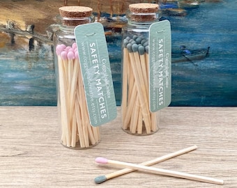 May include: Two glass jars filled with wooden matches. The matches have colored tips, one jar has pink tips and the other has gray tips. The jars have cork stoppers and a label that reads "Coggeshall Creative Safety Matches www.coggeshallcreative.etsy.com".