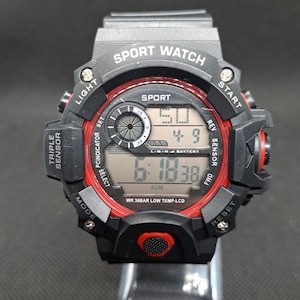 May include: Black and red digital sport watch with a black band. The watch face has a red ring around it with the words "Sport Watch" and "Sport" printed on it. The watch face displays the time, date, and alarm.