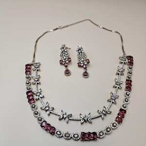 May include: A two-layered necklace and a pair of matching drop earrings featuring deep red oval-cut stones and clear pavé-set crystals. The necklace has a delicate metal chain and a design incorporating floral-shaped crystal accents and red stones.
