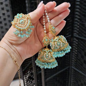 May include: Gold-toned jewelry set featuring a ring and earrings. The ring and earrings have a floral design with turquoise and pearl accents. The earrings are bell-shaped with dangling turquoise beads. The jewelry is displayed against a dark background.