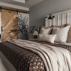 Peut inclure: Un lit doté d'une courtepointe marron foncé à motif floral blanc, surmontée d'un épais jeté en tricot crème. Le lit comprend deux oreillers carrés à motif quadrillé noir, un oreiller rectangulaire texturé et une tête de lit blanche.