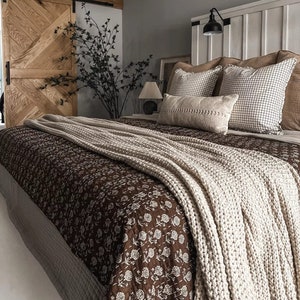 Könnte beinhalten: Schlafzimmer mit einem Bett, das mit einer braunen, floral gemusterten Steppdecke und einer cremefarbenen Strickdecke ausgestattet ist. Dekorative Kissen in verschiedenen Texturen und Mustern sind auf dem Bett angeordnet. Eine Holztür und ein weißes Kopfteil.