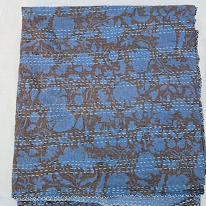 Puede incluir: Una colcha rectangular con una base de color marrón y un patrón floral azul repetitivo. La tela está decorada con filas horizontales de bordado de punto hilván blanco que crean una apariencia acolchada y texturizada en toda la superficie.