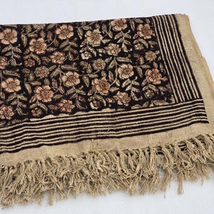May include: A rectangular textile with a floral pattern in shades of brown and beige on a black background. The border features horizontal stripes and fringe. The textile is folded.