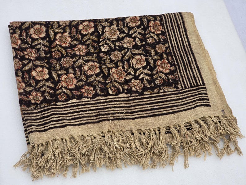 May include: A rectangular textile with a floral pattern in shades of brown and beige on a black background. The border features horizontal stripes and fringe. The textile is folded.