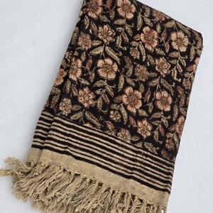 May include: A folded, dark brown textile with a floral pattern in shades of tan and red. The fabric has a striped border with a tan fringe. The textile is likely a scarf, shawl, or throw blanket.