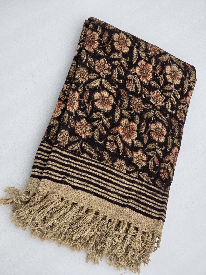 May include: A folded, dark brown textile with a floral pattern in shades of tan and red. The fabric has a striped border with a tan fringe. The textile is likely a scarf, shawl, or throw blanket.