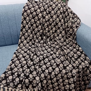 May include: A black and cream floral patterned throw blanket with a fringed edge is draped over a blue sofa. The blanket features a repeating floral design. Yellow flowers in a vase are visible in the background.