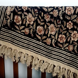May include: A black and beige floral patterned textile with a fringed edge. The textile features a repeating floral design in beige and brown, set against a black background. The border has black and beige stripes.