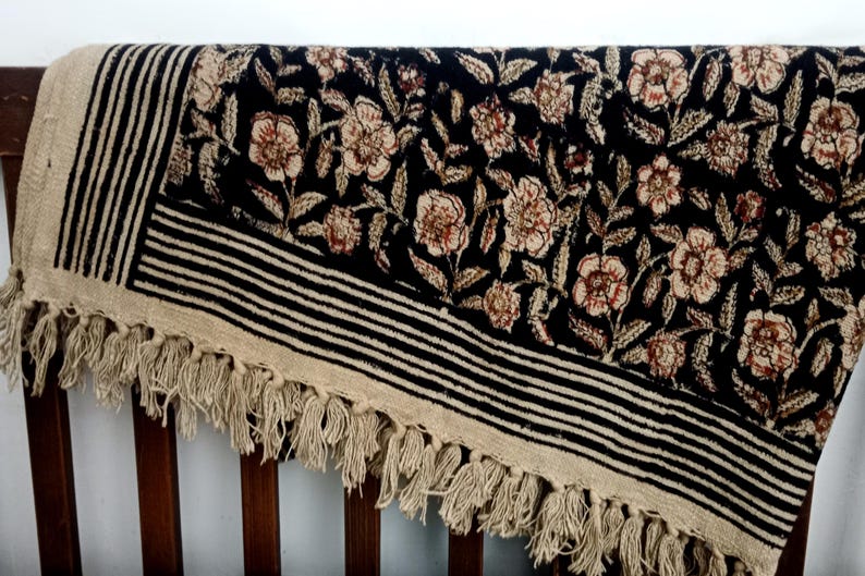 May include: A black and beige floral patterned textile with a fringed edge. The textile features a repeating floral design in beige and brown, set against a black background. The border has black and beige stripes.