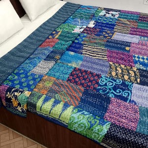Op de afbeelding: Een patchwork quilt in tinten blauw, groen, paars en oranje, met diverse patronen en texturen. De quilt ligt op een bed met witte kussens en een donkerblauwe rand. Waarschijnlijk handgemaakt.